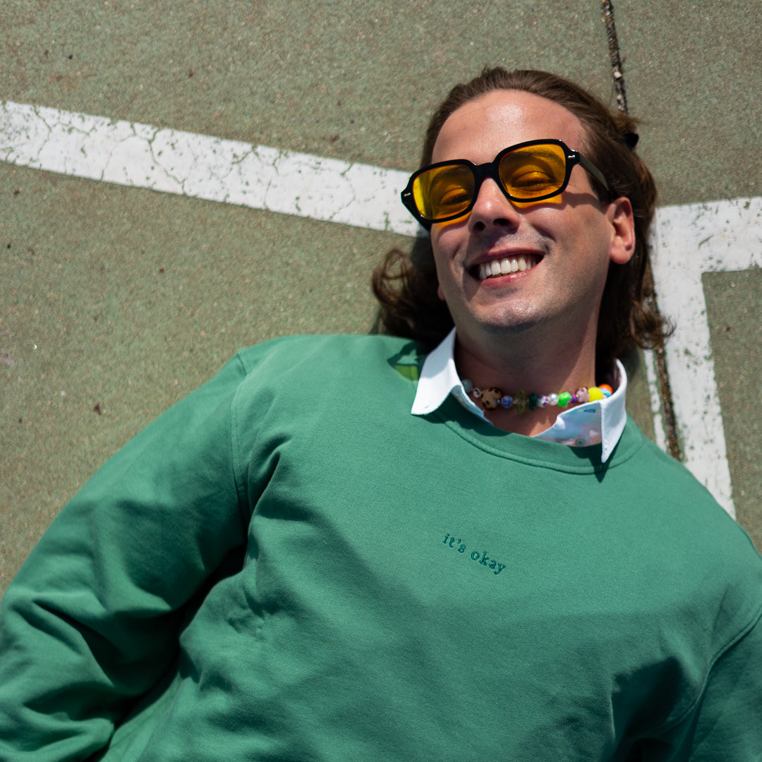 it's okay crewneck: green made in portugal with organic cotton. Join us. Image: boy on the floor wearing sunglasses and a necklace smiling