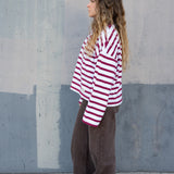 Person wearing a red and white striped shirt and brown pants standing against a gray wall (side)