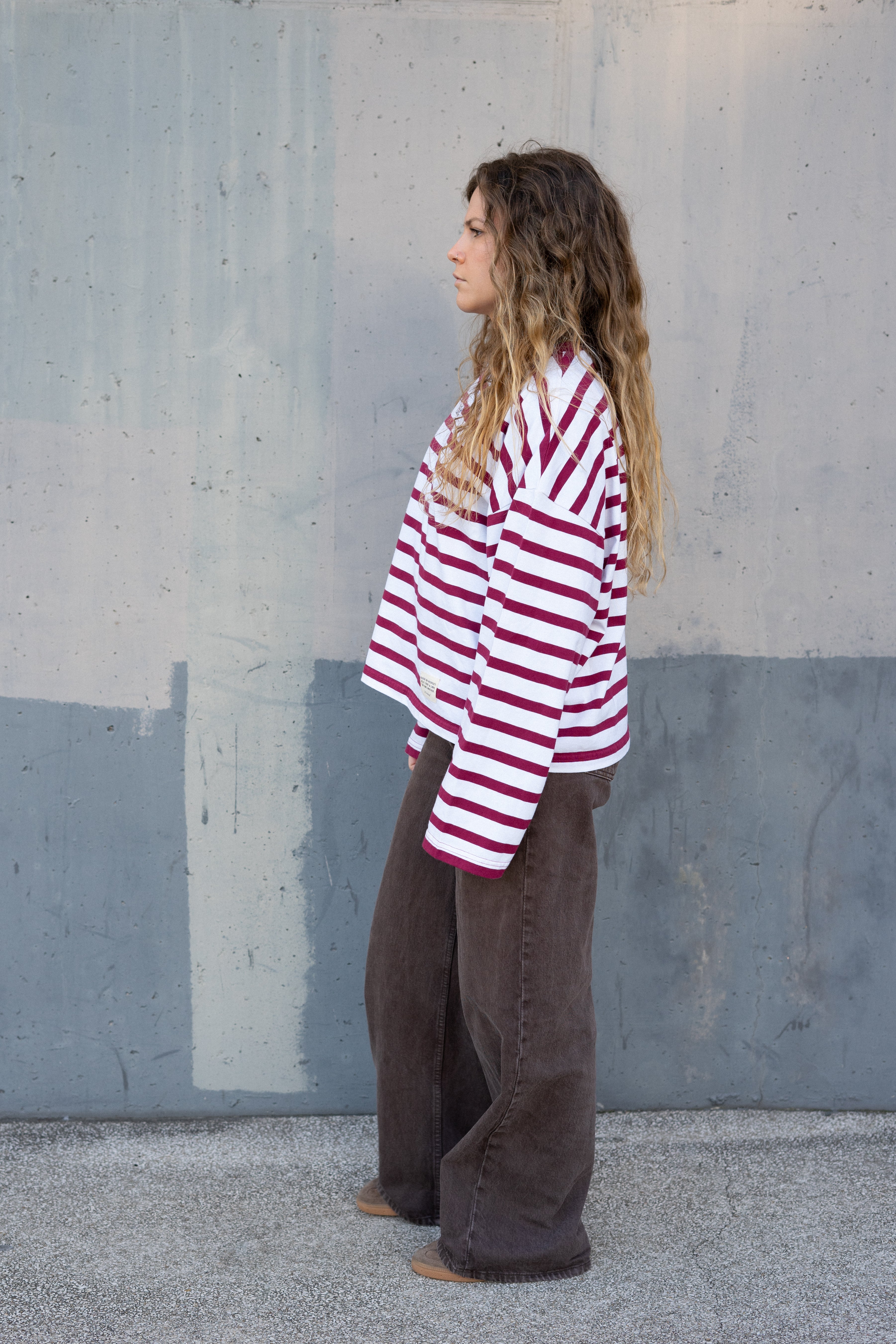 Person wearing a red and white striped shirt and brown pants standing against a gray wall (side)