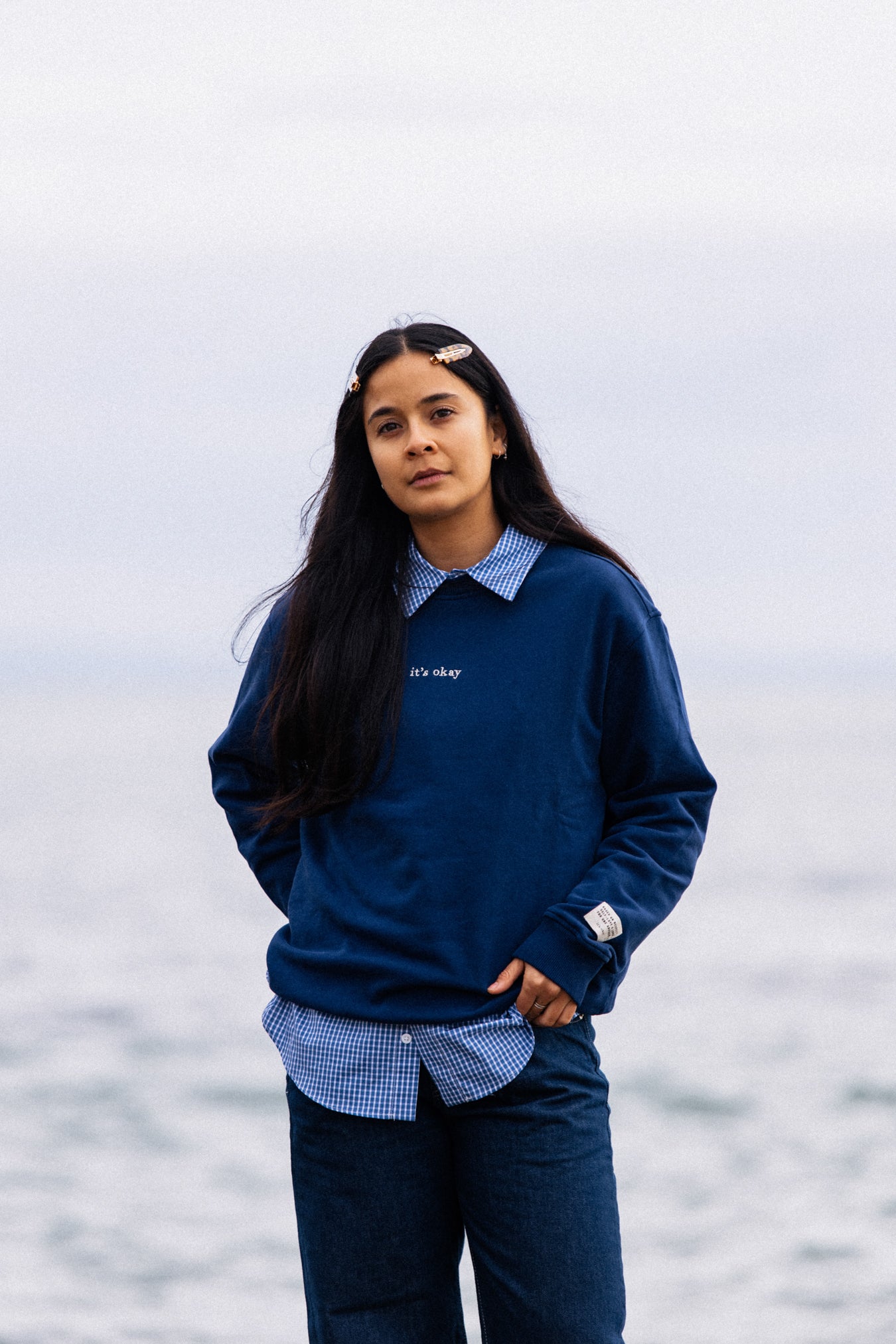 Woman wearing navy IT’S OKAY organic cotton crewneck outdoors by the sea, relaxed fit.