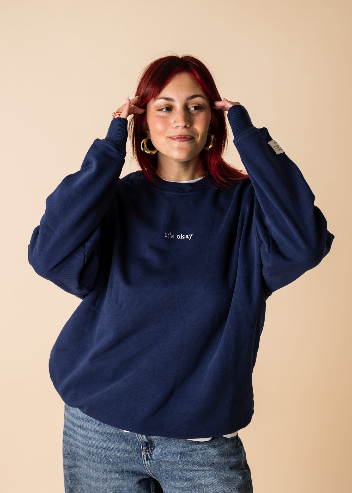 A person wearing a blue crewneck sweatshirt with embroidery on the front and back, posing with one hand raised to their head.