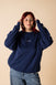 A person wearing a blue crewneck sweatshirt with embroidery on the front and back, posing with one hand raised to their head.