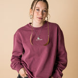 A person wearing a purple crewneck sweatshirt with a front and back embroidery, standing against a beige background.