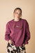 A person wearing a purple crewneck sweatshirt with a front and back embroidery, standing against a beige background.