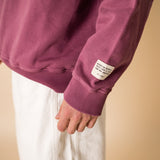 Close-up of burgundy IT’S OKAY organic cotton crewneck sleeve showing woven label detail.