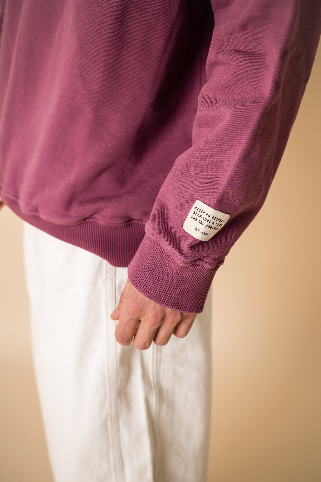 Close-up of burgundy IT’S OKAY organic cotton crewneck sleeve showing woven label detail.