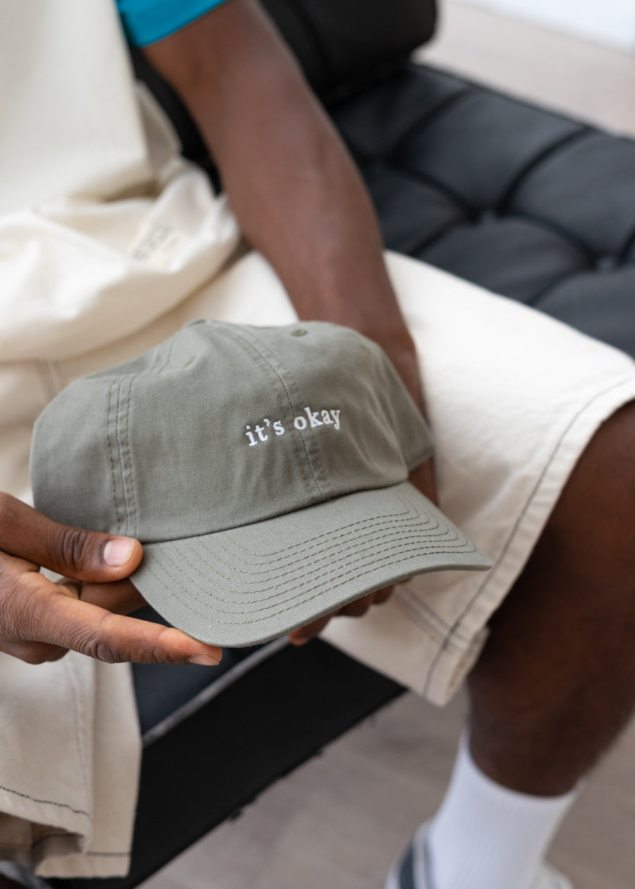 it's okay organic cap green | made with organic cotton, embroidered. The hat is on a black men's hands, he is wearing white shorts. 