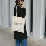 it's okay tote bag: organic cotton with black ink. Photo: girl with long coat and blackberry it's okay cap wearing the tote bag