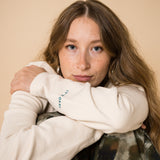 Blond girl wearing an organic crew neck sweater in off-white, seamless design. Relaxed and minimal style, perfect for any occasion. Studio Photography gender neutral apparel. close up of the face and embroidery: Cultivate what matters