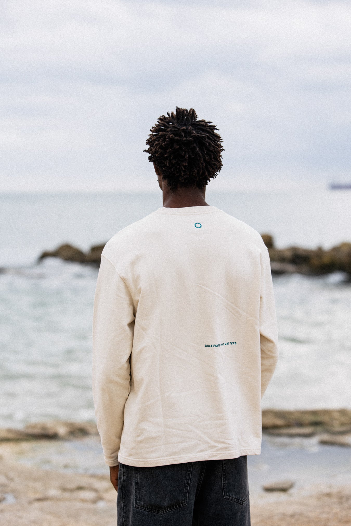Model José wearing Cultivate Crew at the beach. Photo of the back of the crewneck: cultivate what matters